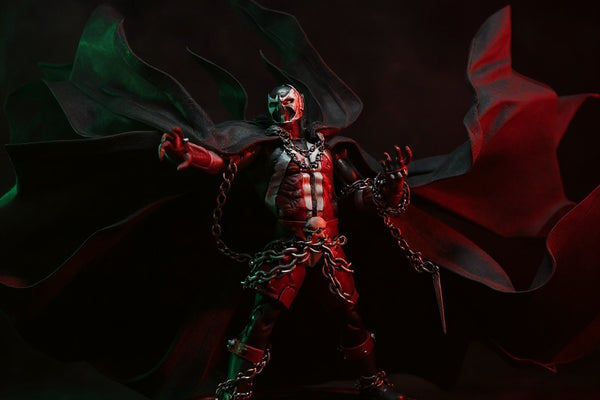 Black fabric wire-frame cape with flame-like torn edges draped over a Spawn-style action figure torso, displayed on a pedestal against a gothic fog backdrop with red spotlight.