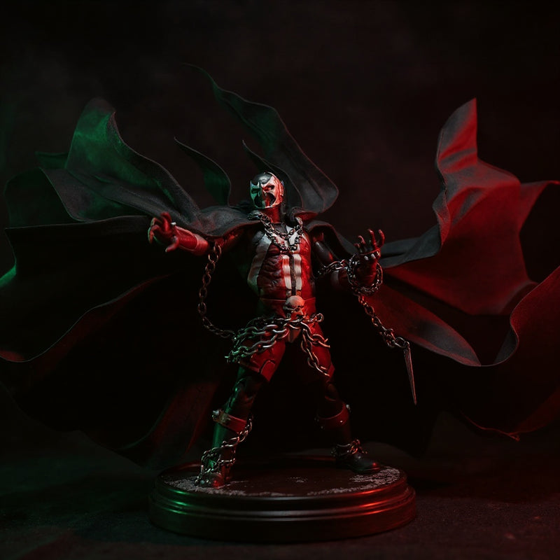Black fabric wire-frame cape with flame-like torn edges draped over a Spawn-style action figure torso, displayed on a pedestal against a gothic fog backdrop with red spotlight.