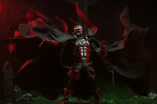 Black fabric cape with flame-like torn edges draped over a Spawn-style action figure on a pedestal, lit by red spotlight in a gothic backdrop with fog and chains.
