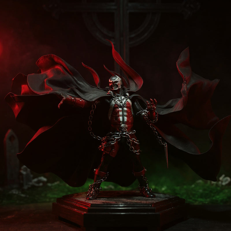 Black fabric cape with flame-like torn edges draped over a Spawn-style action figure on a pedestal, lit by red spotlight in a gothic backdrop with fog and chains.
