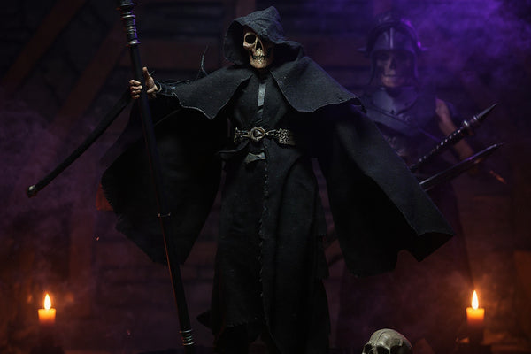 Dark textured fabric cape with visible seams and wire-enhanced drape flowing over a black display platform, surrounded by miniature skulls and bones, fog, and candlelight against a deep violet and blue gradient background.