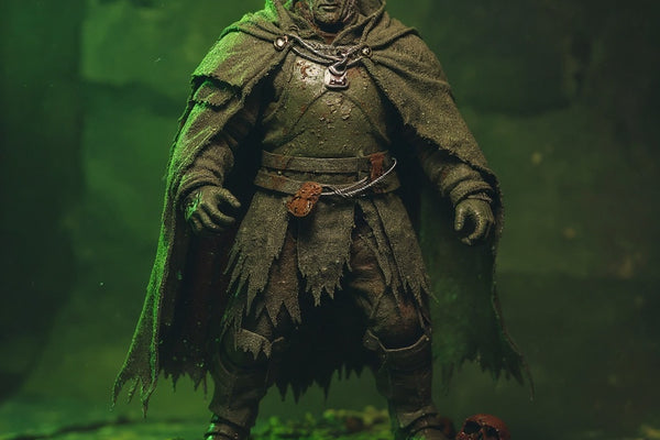 A weathered grey-green fabric cape draped over a 1/18 scale armored action figure shoulder, set in a battle-damaged diorama with rusted metal debris, skull fragments, and green fog lighting.