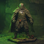 A weathered grey-green fabric cape draped over a 1/18 scale armored action figure shoulder, set in a battle-damaged diorama with rusted metal debris, skull fragments, and green fog lighting.