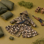 Miniature camo tactical coat and pants set laid flat on a green-to-tan gradient background surrounded by military diorama props.