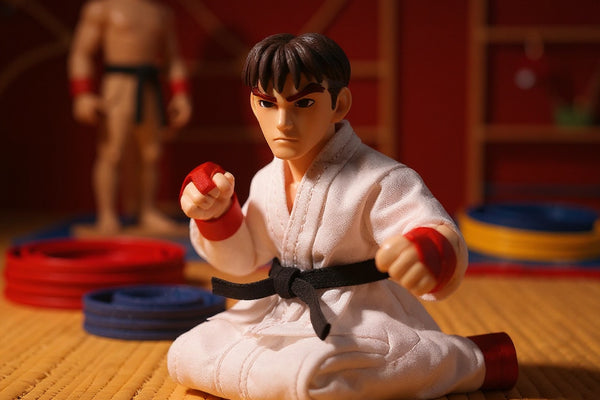 Small fabric karate gi with visible stitching laid flat on a bold red-orange gradient background, surrounded by miniature wooden training dummy and colorful folded karate belts.
