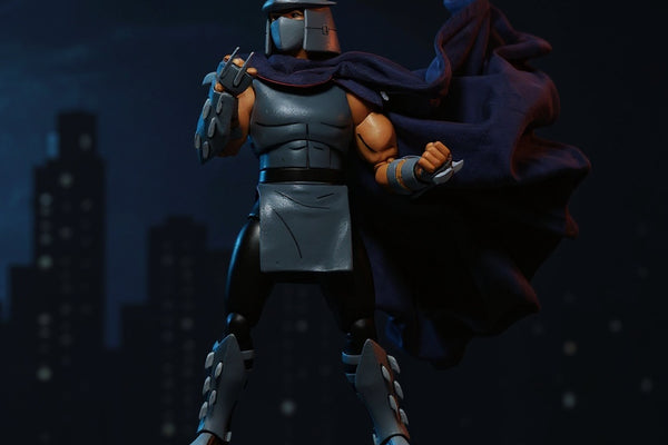 Black fabric cape with red trim on Shredder figure, posed on a rooftop diorama with neon city backdrop and dramatic lighting.