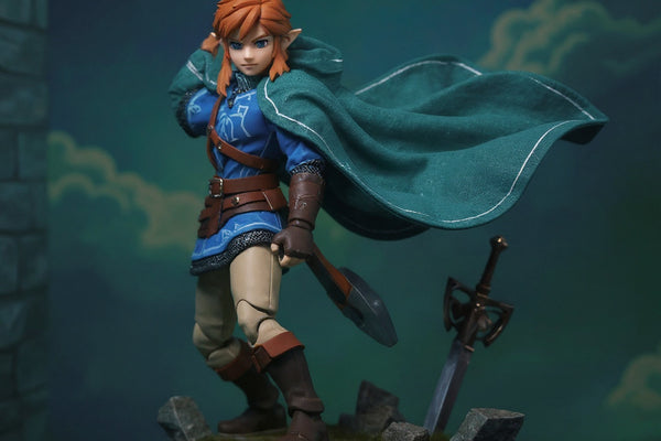 Flowing blue-green fabric cape with wire-shaped folds draped over miniature armor torso on Hyrule-themed diorama with rock formations, wind fan, Master Sword, and blue-hour lighting