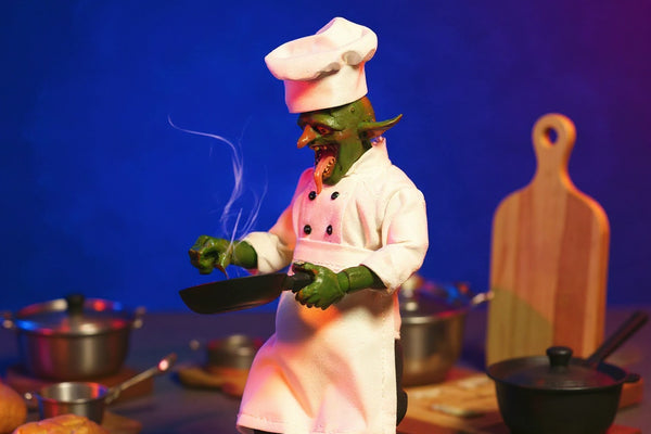 Miniature chef costume laid flat on a miniature kitchen counter with tiny utensils and food props, bold blue and magenta gradient background.