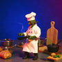 Miniature chef costume laid flat on a miniature kitchen counter with tiny utensils and food props, bold blue and magenta gradient background.