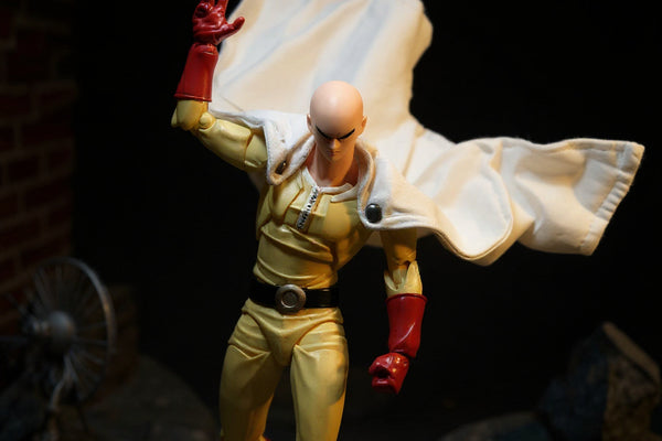Yellow-and-white fabric action figure cape billowing on a mannequin torso against a bold rooftop diorama with dramatic lighting