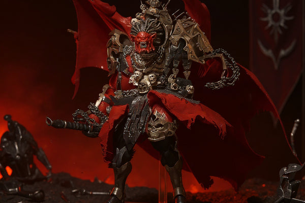 Dark crimson-red and bronze textured cloth cape with visible folds draped on a small display stand, miniature battlefield backdrop with weapons and chaos banner, red and black ambient lighting.